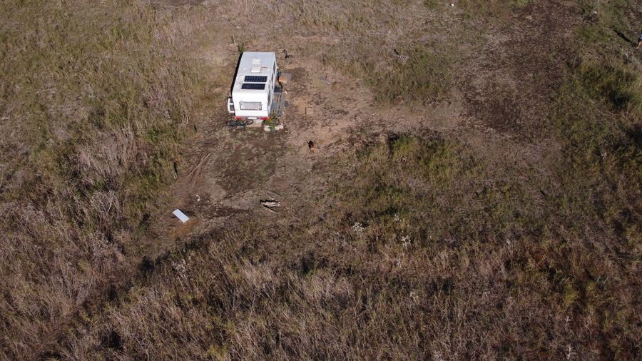 Aerial photo taken by a drone over dry scrubland with a caravan; a small brown dog stands in the middle of the clearing